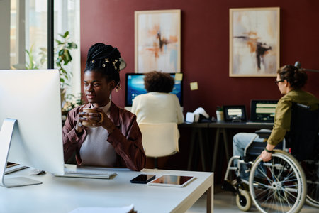 Young female designer with cup of coffee looking at computer screen while sitting by workplace and reading task pointsの写真素材