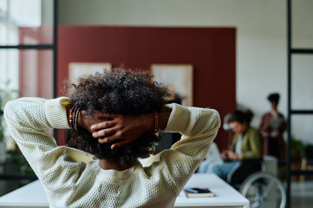 Rear view of young businessman keeping hands on back of head while sitting by workplace in front of camera and looking at colleaguesの写真素材