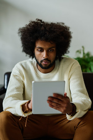 Young serious male designer in white sweater looking at tablet screen while sitting in front of camera and carrying out AI design taskの写真素材