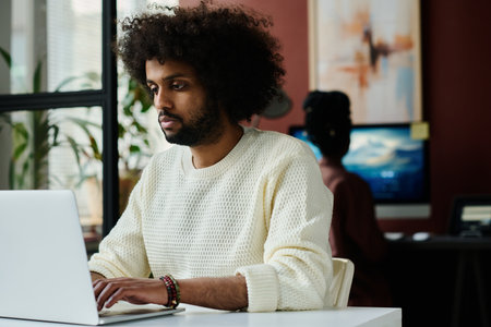Young serious male designer in white sweater looking at laptop screen and pressing keys of keyboard...