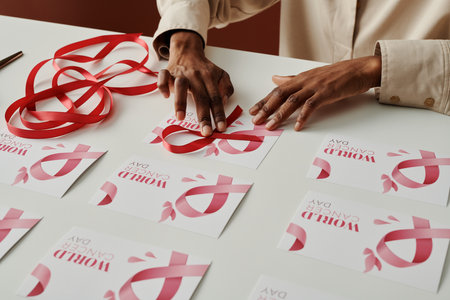 Hands of young unrecognizable black woman putting pink ribbon on world cancer day postcard while preparing for prevention campaignの写真素材