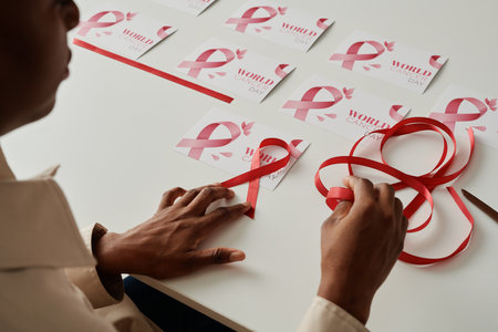 Hands of young woman with pink ribbon making symbols of breast cancer and fixing them on handmade postcards while sitting by deskの写真素材