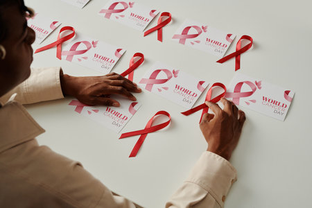 Close-up of African American girl sitting by desk with group of world cancer day postcards and putting pink ribbons on themの写真素材