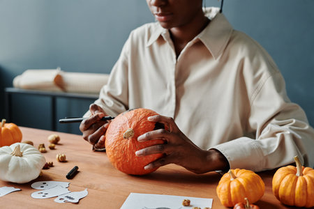 Cropped shot of young African American woman with highlighter and ripe pumpkin sitting by desk and preparing for Halloweenの写真素材