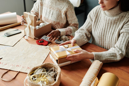 Cropped shot of young woman in woolen sweater keeping hands on stack of packed Christmas gifts while sitting next to her friendの写真素材