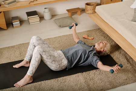 High angle of aged woman in activewear lying on mat in bedroom and doing exercise for arm and chest muscles during morning workoutの写真素材