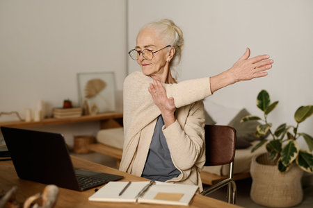 Senior woman in eyeglasses looking at screen of laptop and doing exercise for stretching while sitting by table and working with online dataの写真素材