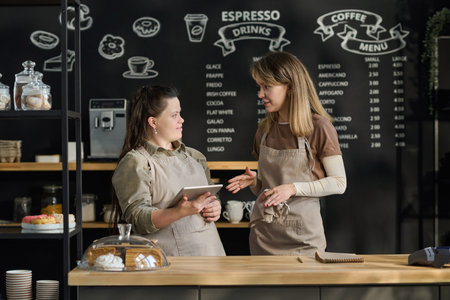 Young blond waitress explaining something to female colleague with disability during discussion by counter against board with menuの写真素材