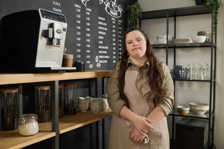 Young woman with Down syndrome wearing apron looking at camera while standing by coffee machine against blackboard with menuの写真素材