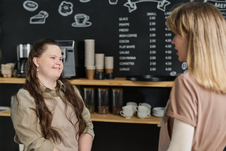 Happy young waitress with Down syndrome and her colleague having chat while standing by workplace against blackboard with menuの写真素材