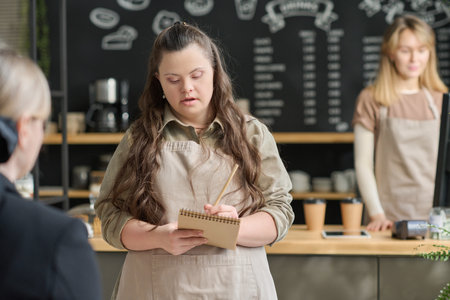 Young woman with disability writing down order of client in notepad against her colleague standing by counter in front of computerの写真素材