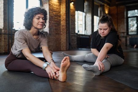 Girl with Down syndrome repeating after young female fitness trainer while both sitting on mats in gym during physical exerciseの写真素材