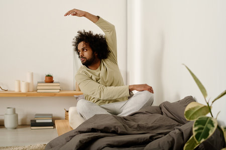 Young multiethnic man in pajamas stretching arm during side bend exercise while sitting on bed in the morning after sleepの写真素材