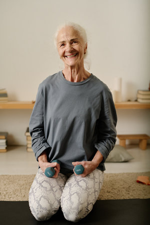 Happy elderly woman in cotton leggins and pullover holding dumbbells while standing on knees on the floor and looking at cameraの写真素材