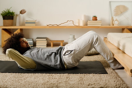 Active young man in sweatpants and hoodie doing sit ups while lying on black mat during morning...