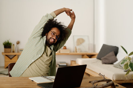 Young man in casualwear raising stretched arms over head and bending aside while sitting in front of laptop and enjoying minute of restの写真素材