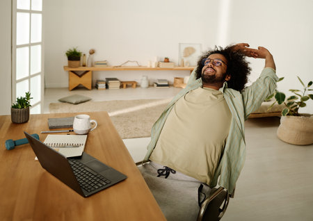 Lazy young male student in casualwear stretching backwards while sitting on chair in front of laptop and looking through online informationの写真素材