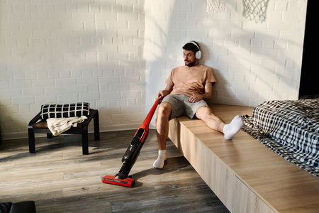 Young relaxed man in headphones sitting against wall of bedroom and listening to music while cleaning floor with vacuum cleanerの写真素材