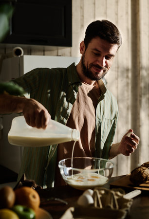 Young man pouring fresh milk from plastic bottle into glass bowl with broken fresh raw eggs while standing by kitchen counterの写真素材