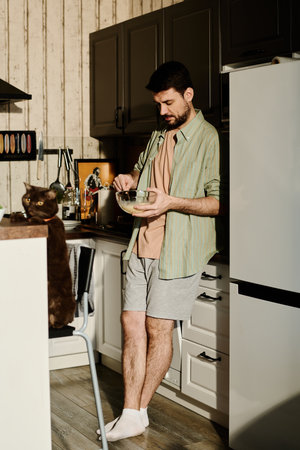 Young man in casualwear standing by refrigerator and beating raw eggs with fresh milk in glass bowl while preparing breakfastの写真素材