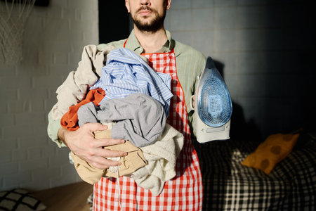Cropped shot of young bearded man holding electric iron and pile of clean crumpled clothes while...