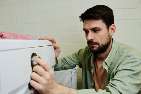 Serious young man squatting by automatic washing machine in bathroom and choosing settings before pressing start buttonの写真素材