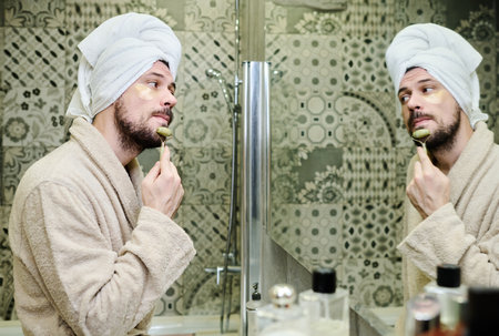 Young man in bathrobe having procedure of facial massage with jade stone roller after morning shower...