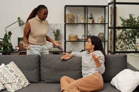 Angry young African American woman shouting at her confused girlfriend sitting on couch and trying to understand what happenedの写真素材