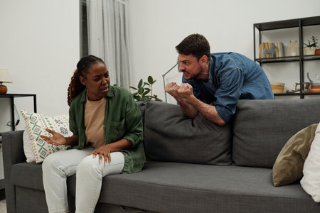 Young furious man showing punches while bending over couch and looking at his wife with angry expression during quarrelの写真素材