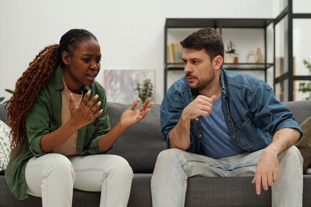 Young African American woman trying to explain something to her husband during argument while both sitting on couchの写真素材