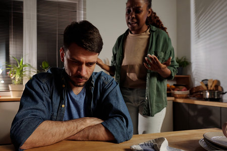 Young stressed man with crossed arms by chest sitting by kitchen table while his irritated wife shouting at him during argumentの写真素材