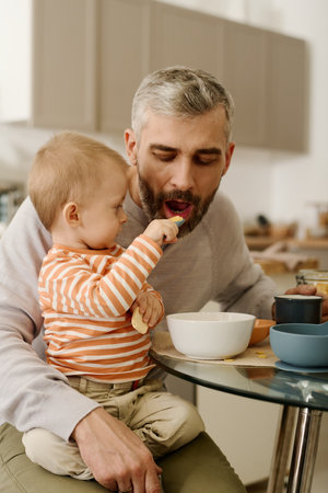 Adorable baby boy putting spoon with cornflakes into father mouth while sitting on his knees by small table with bowls during breakfastの写真素材