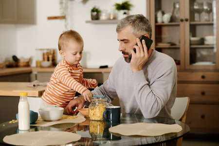 Mature businessman with mobile phone by ear sitting by kitchen table next to his cute baby son taking cornflakes from open jarの写真素材