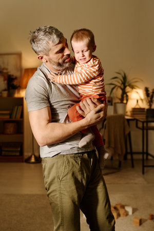 Mature man in grey t-shirt and beige pants holding crying naughty baby son on hands and looking at...