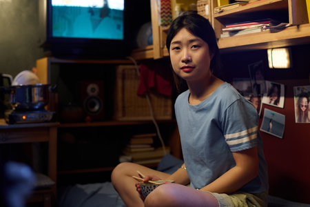 Young Asian woman with chopsticks and empty bowl in hands sitting on bed in small apartment and looking at camera after lunchの写真素材