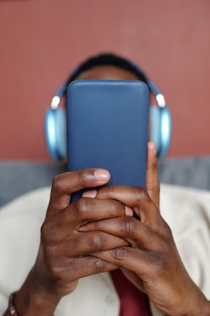 Smartphone in hands of young unrecognizable African American man in headphones relaxing in front of...