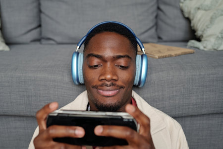 Young male gamer in headphones sitting in front of camera against couch and looking at screen of...