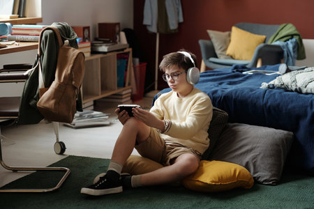 Cute schoolchild in headphones sitting on cushions on the floor of his bedroom and looking at phone screen in gamepadの写真素材