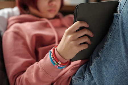 Hand of unrecognizable teenage girl in pink hoodie holding tablet while sitting in front of camera...