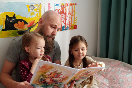 Young bearded man discussing pictures in books of comics with his two adorable daughters while...