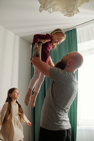 Happy young man holding his adorable laughing girl on hands and looking at her while playing and having fun with his daughtersの写真素材