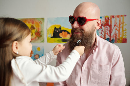 Little girl playing with thick beard of her happy bald father in heartshaped sunglasses and shirt standing in front of his daughterの写真素材