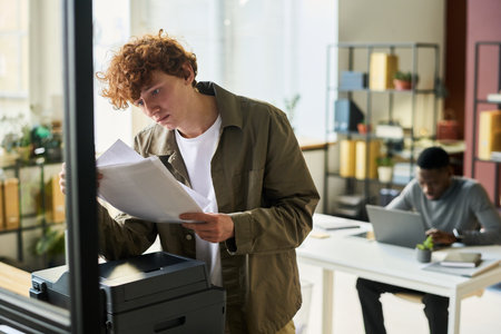 Young male white collar worker in casualwear looking at papers in his hands while standing in front...