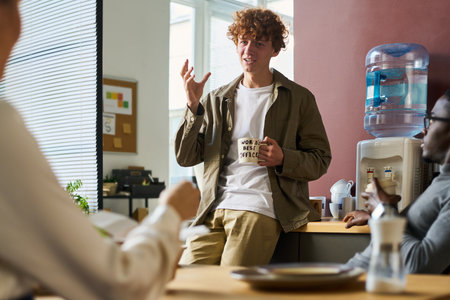 Guy with cup of coffee in casualwear standing by counter with water cooler in front of colleagues and telling them curious story at breakの写真素材