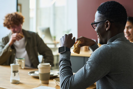 Side view of young African American male office worker with snack sitting by table in front of colleagues and enjoying lunch and chatの写真素材