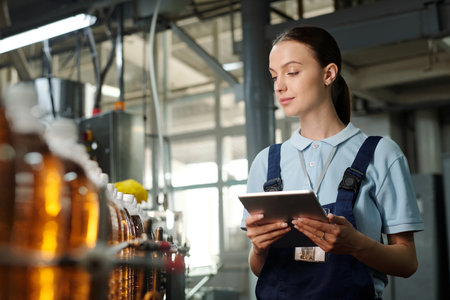 Young female inspector with tablet looking at assembly line with capped bottles with drinks and...