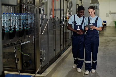 Young female and male engineers of quality control in workwear looking at screen of tablet while...