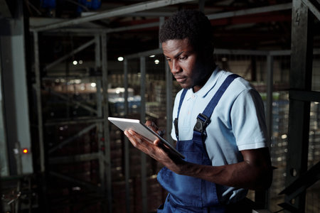 Young male engineer of factory warehouse with tablet scrolling through data on screen while standing...