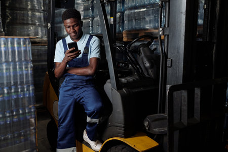 Young male engineer of industrial plant sitting by door of forklift and texting in mobile phone...