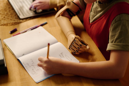 Hand prosthesis of youthful diligent schoolgirl making notes in copybook while sitting by desk and...